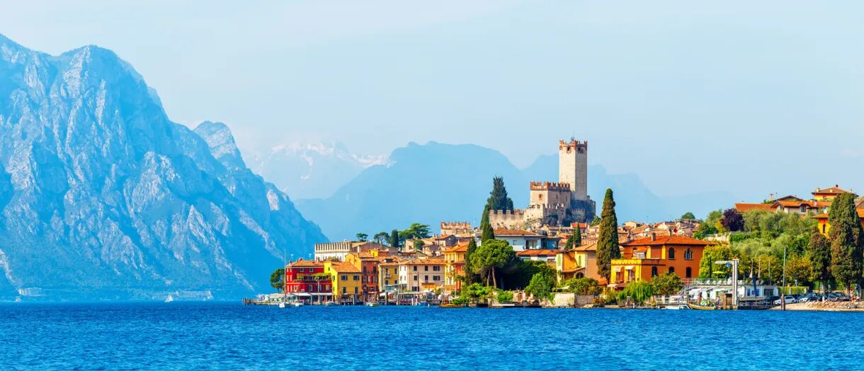 Blick auf den Gardasee mit bunten Häusern, einer mittelalterlichen Burg und den umliegenden Bergen.